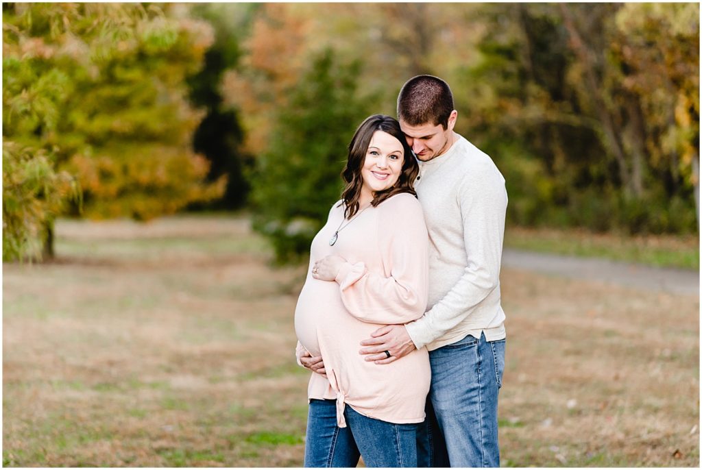 expecting parents cuddling posing in park holding belly