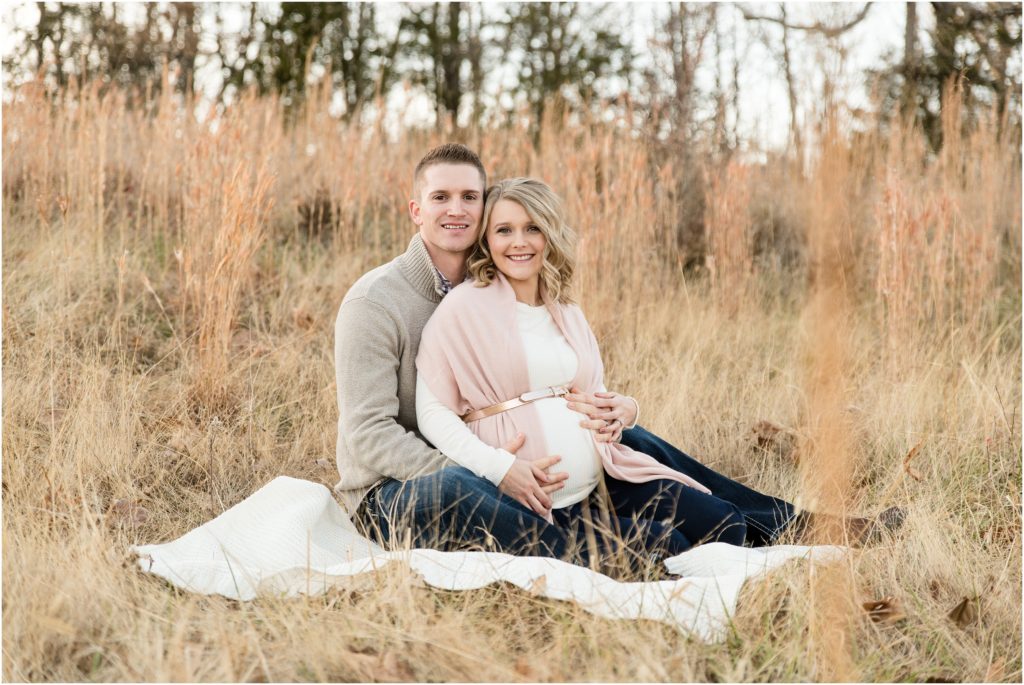couple sitting on blanket holding baby belly for maternity session in tall grass