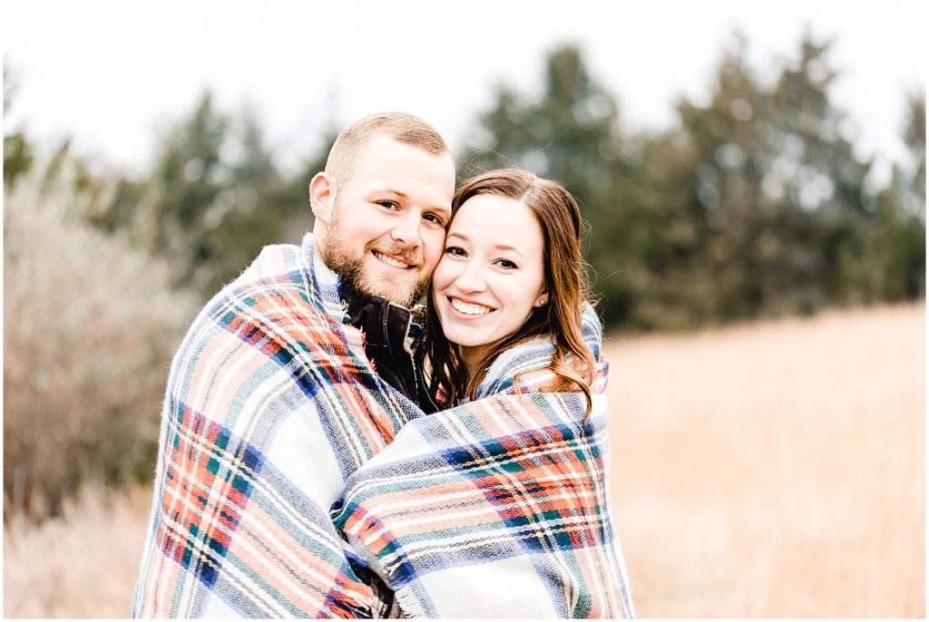 a young couple wrapped up in a plaid blanket smiling at the camera