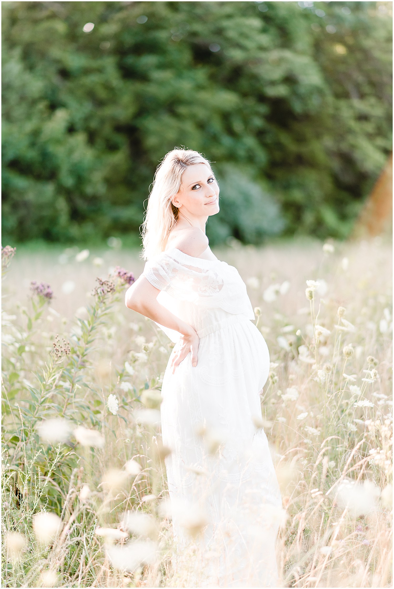 pregnant mom posing hand on back and belly long white dress wildflower fields smiling at camera