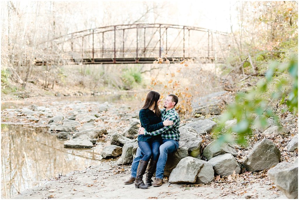 couple sitting on rocks in creek bed smiling at each other capen park bridge engagement session