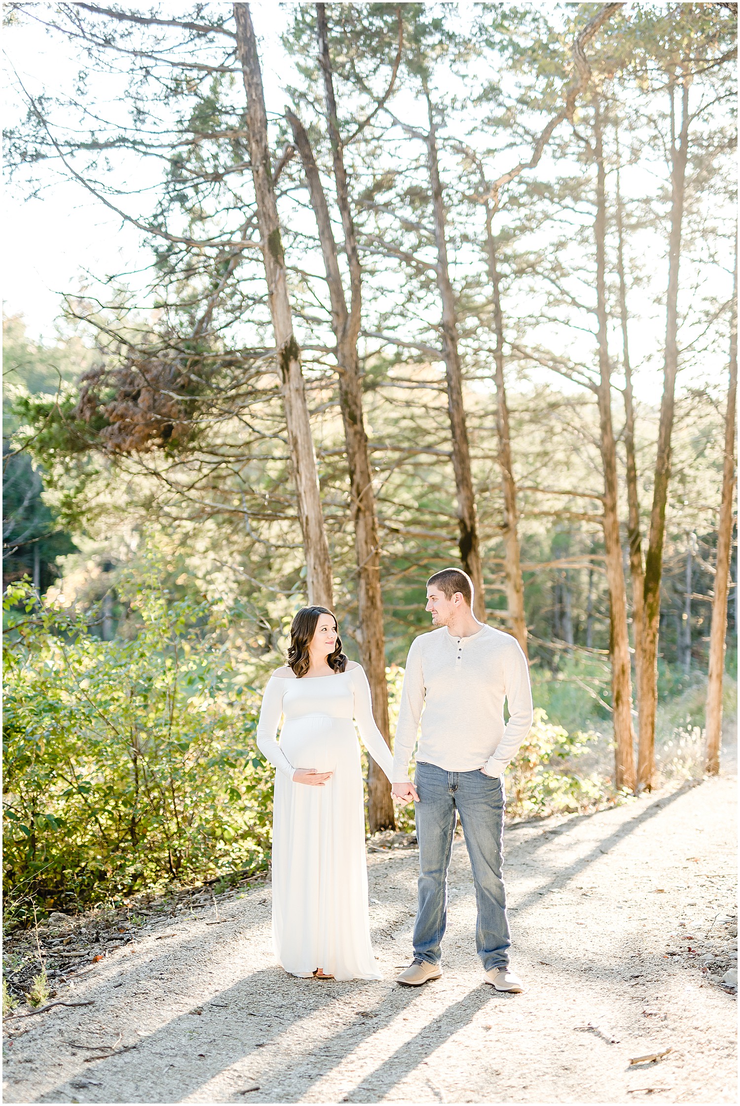 expecting couple standing under pine trees on path sunlight maternity session
