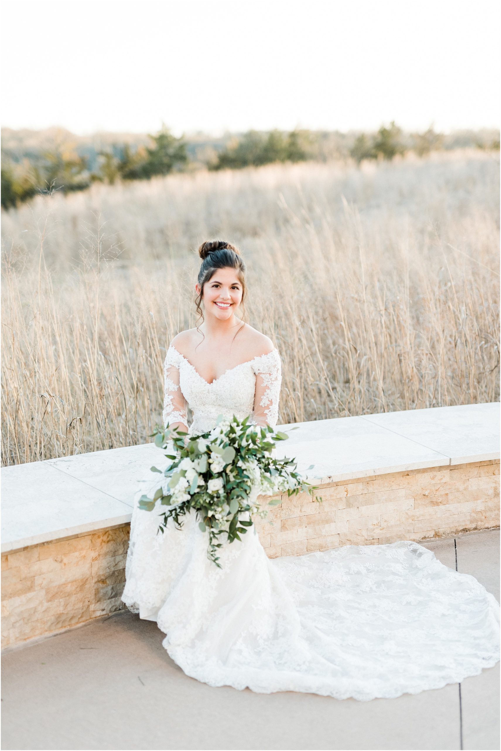 bride with brown bun and long sleeved wedding dress sitting on white bench with green and white bouquet