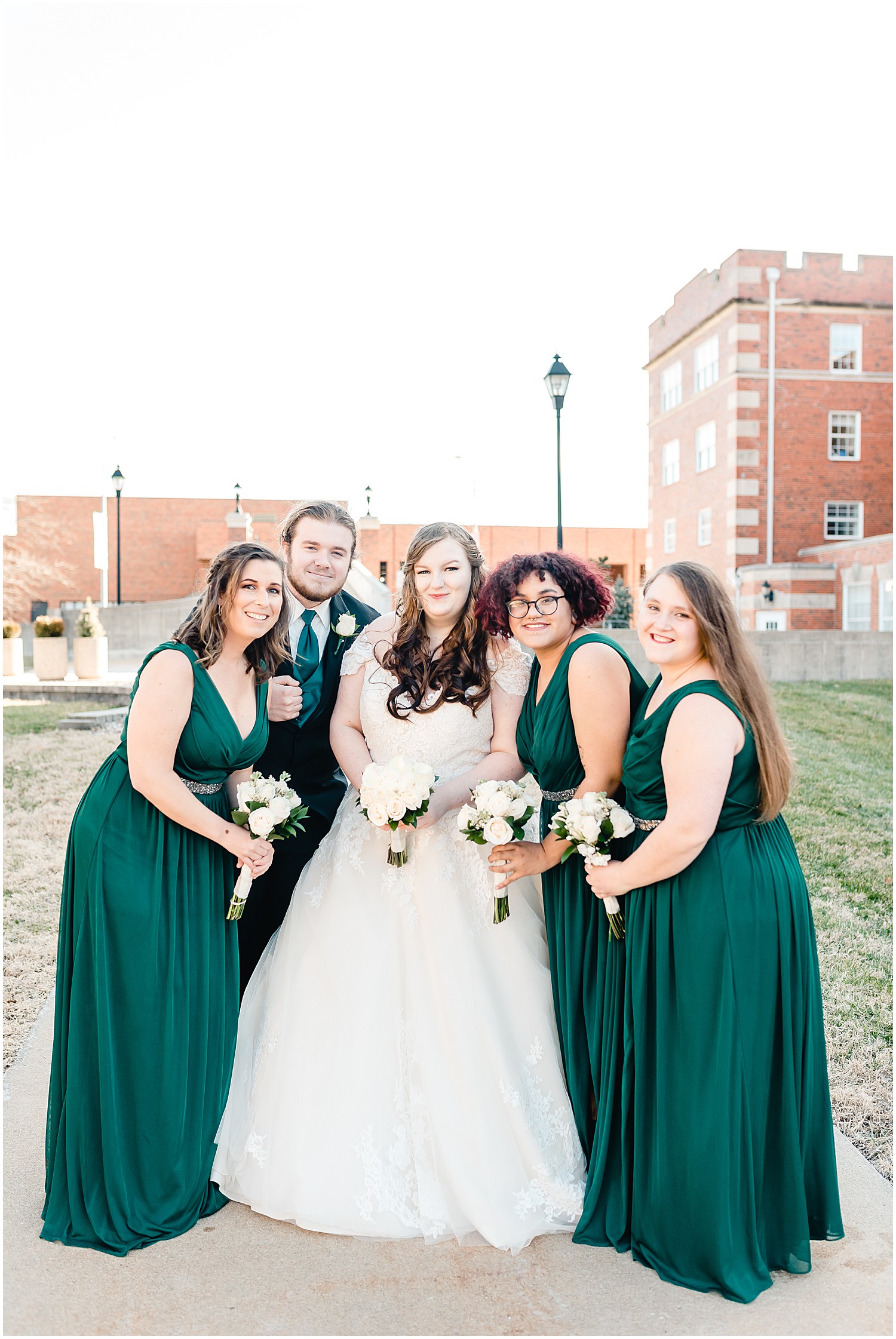bride with her bridal party in emerald dresses and white rose bouquet