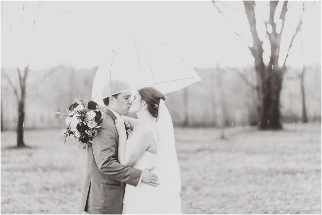 black and white portrait of bride and groom kissing in rain under a clear umbrella