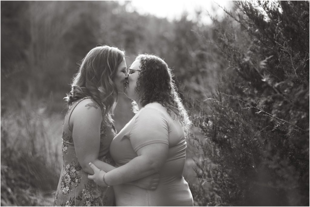 black and white image of engaged female couple kissing by cedar tree