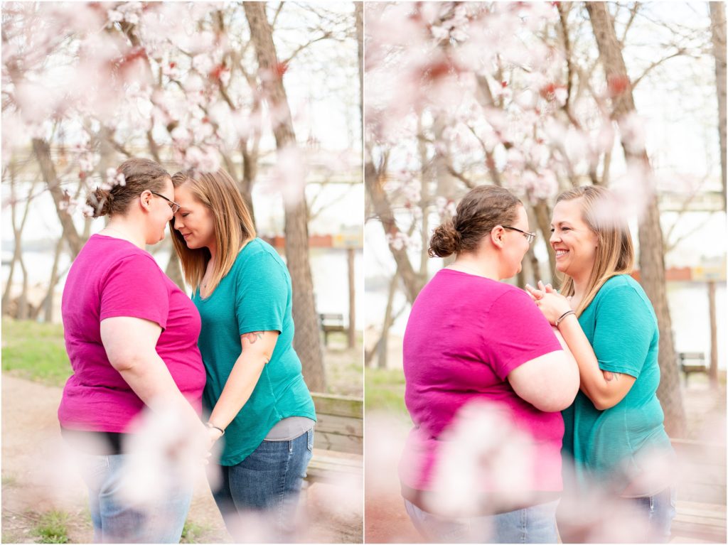 lesbian couple standing and smiling under cherry blossom tree in front of Missouri River for engagement session