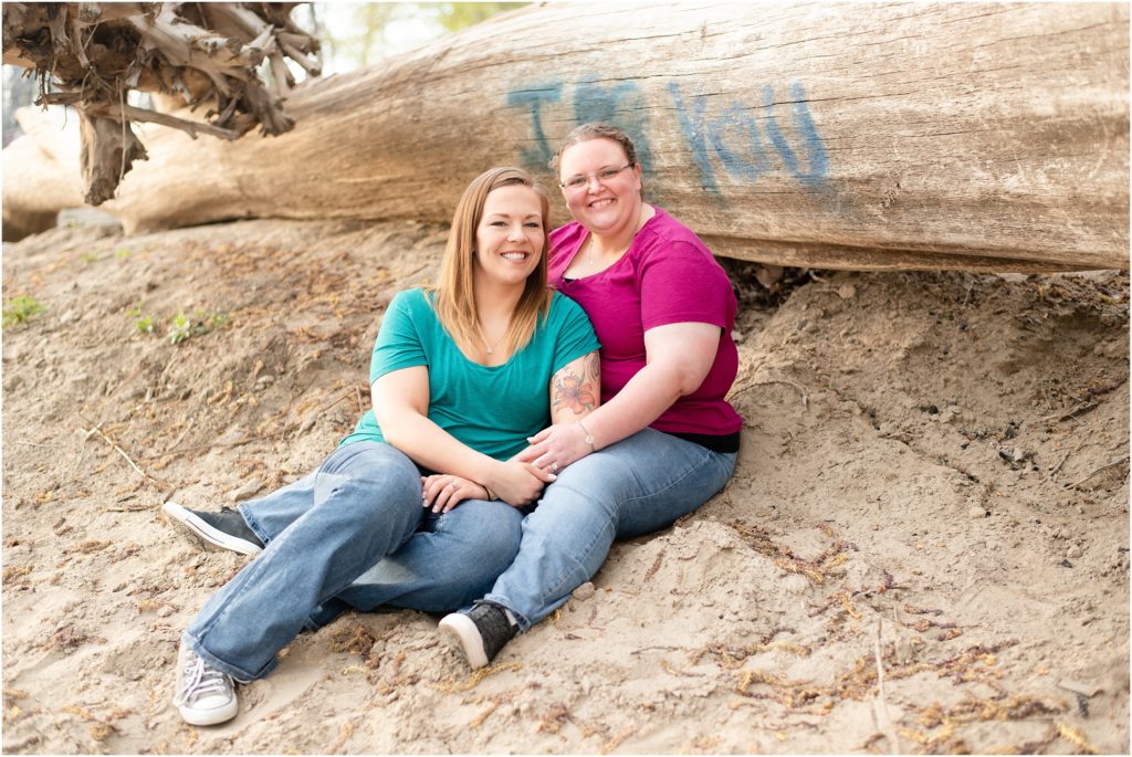 engaged female couple sitting on sand at noren river access by tree that says I love you for engagement session