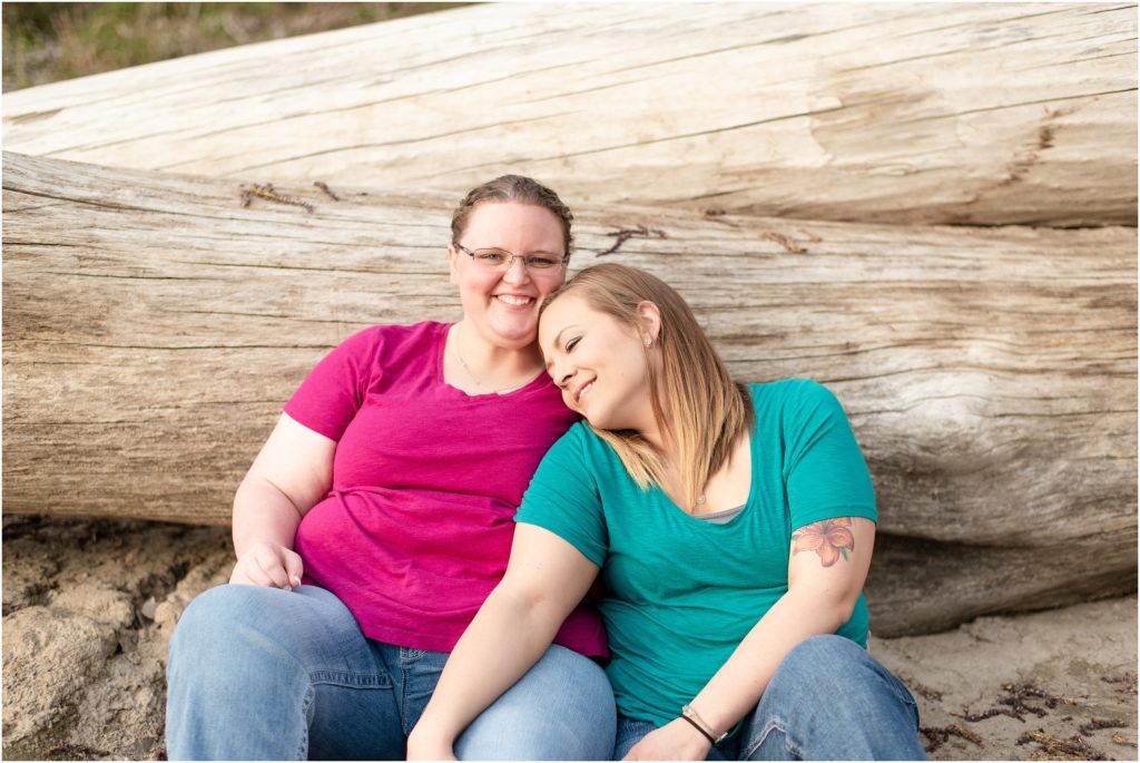lesbian couple wearing blue and pink sitting against fallen tree at noren river access engagement session