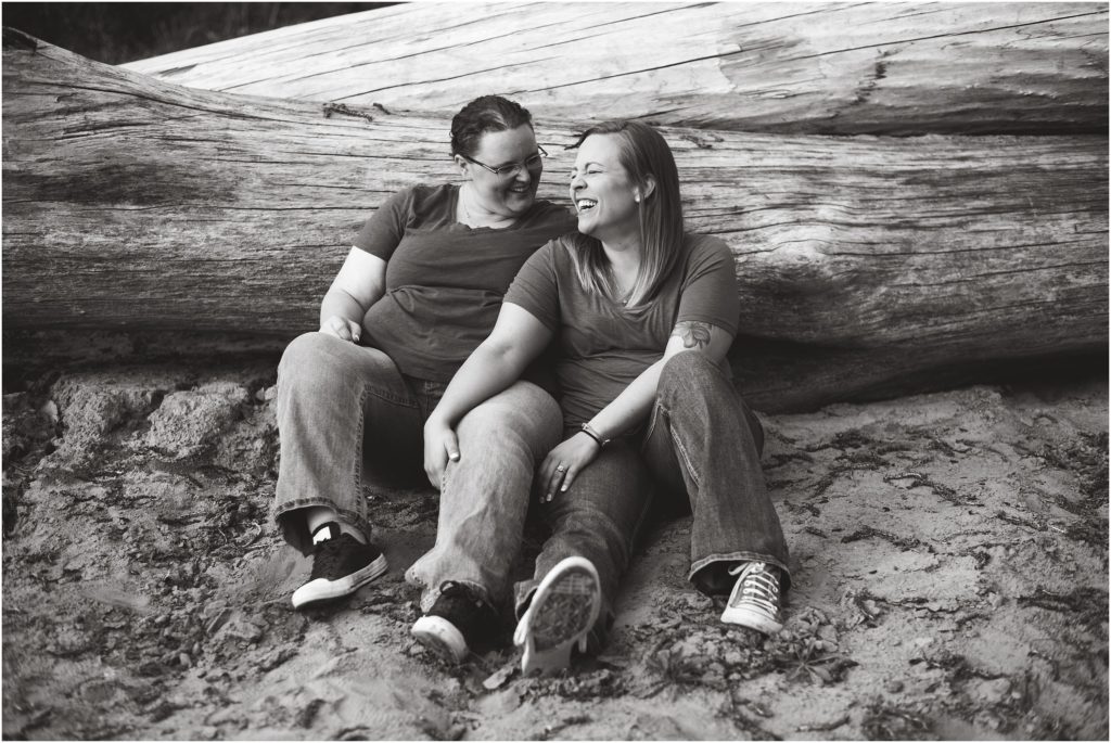black and white image of lesbian couple sitting on sand by fallen tree smiling for engagement photos in Jefferson City mo engagement session