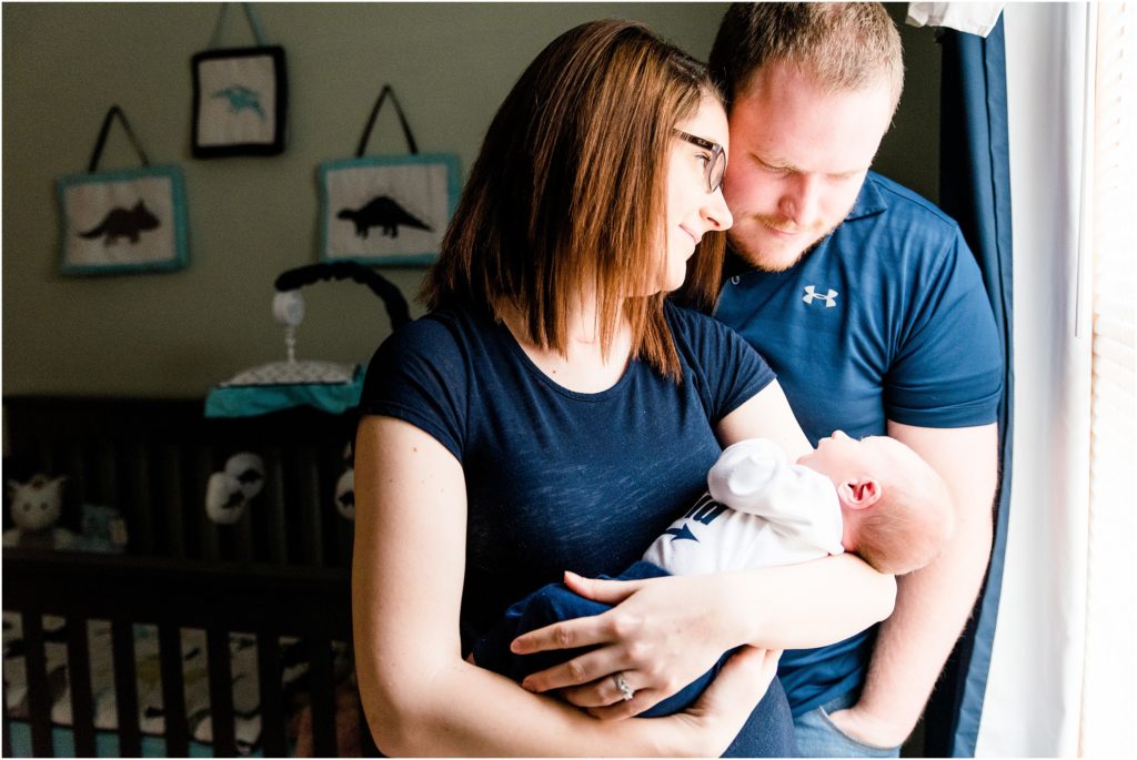 mom and dad holding newborn boy in front of window in navy shirts