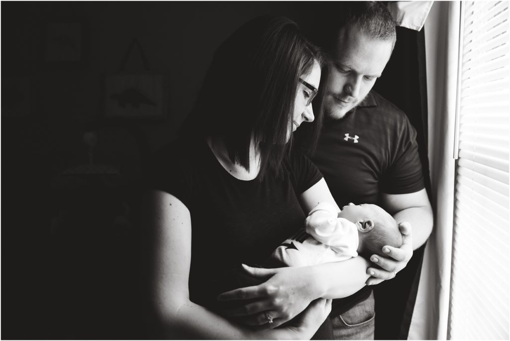 black and white image of mom and dad holding newborn boy in front of window