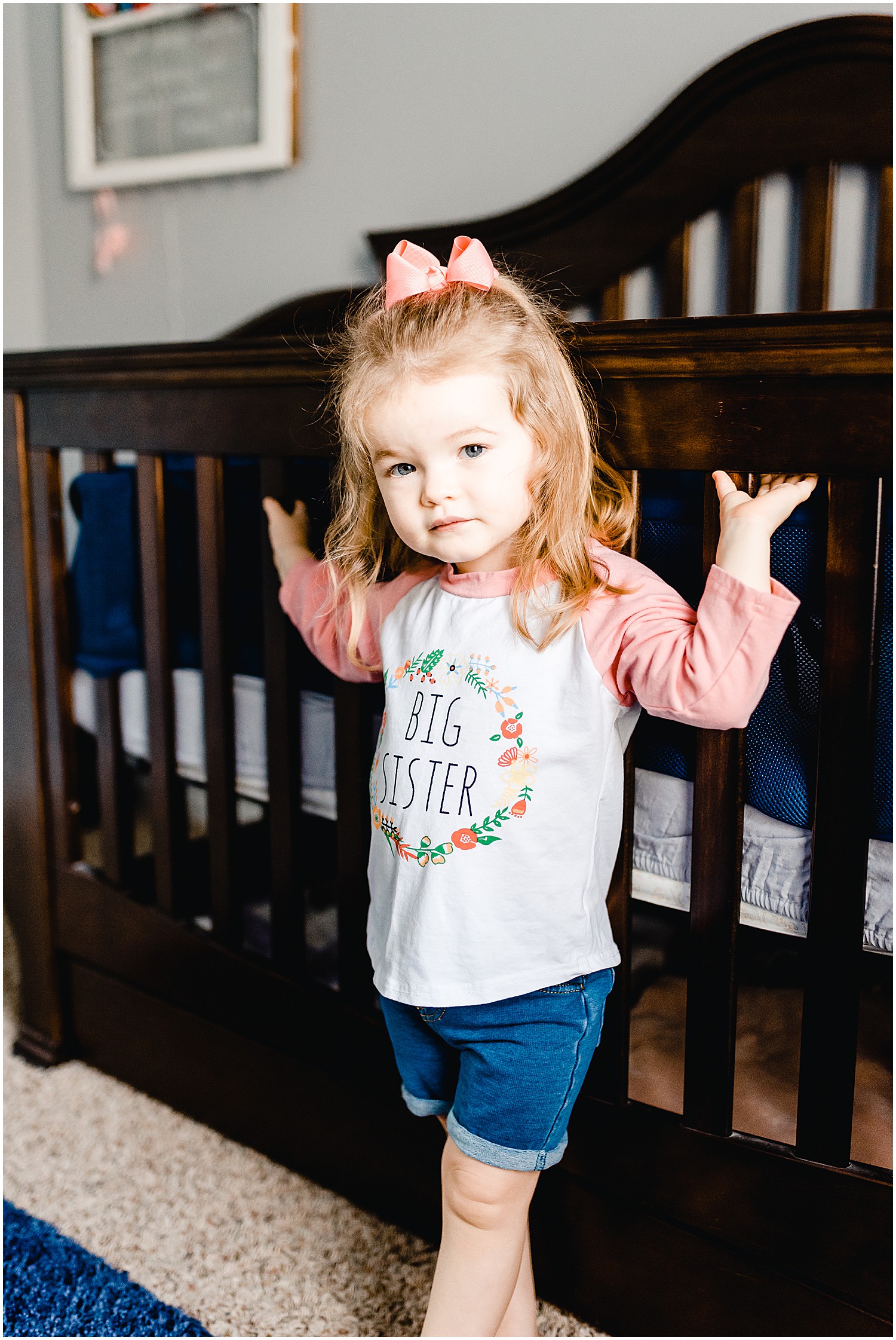 little girl wearing big sister shirt looking at camera next to crib
