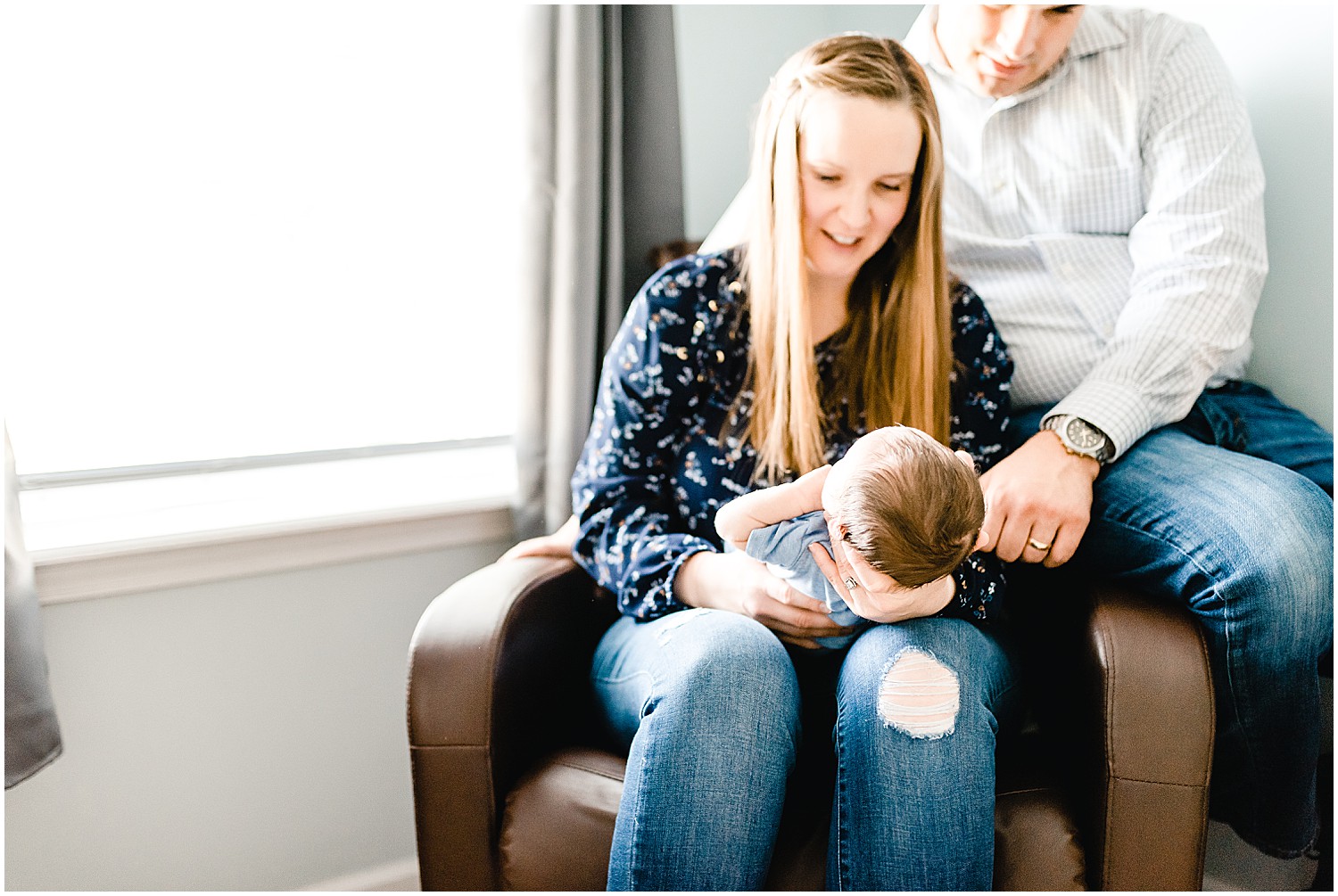 parents sitting on chair holding baby boy next to window for newborn session