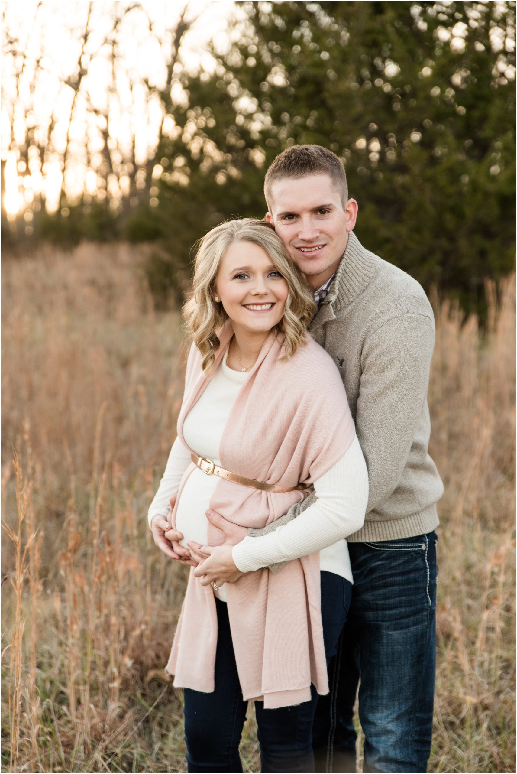 couple smiling at camera and holding baby belly for binder park maternity session in tall grass and sunset