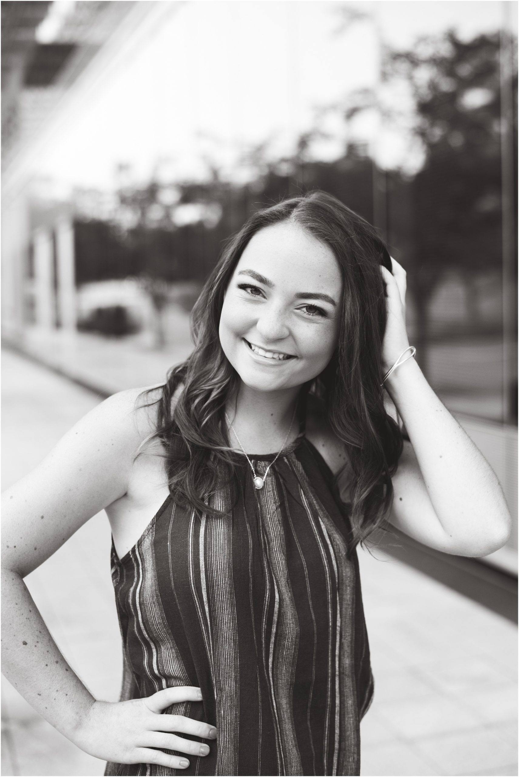 black and white image of senior girl smiling at camera with hand in hair outside of glass building for senior session