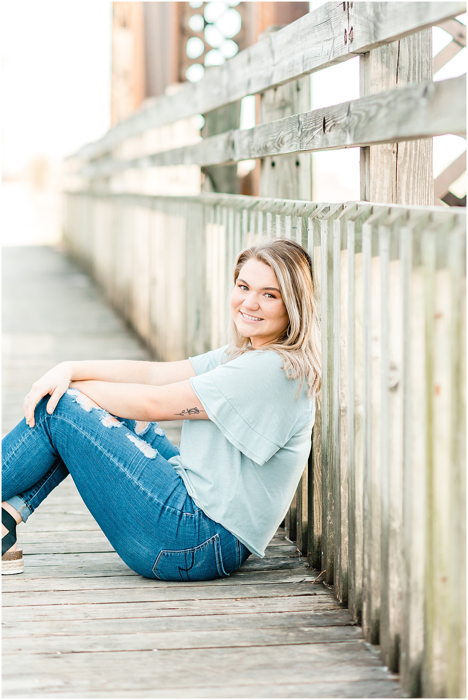 senior girl posing in blue shirt and blue jeans sitting on katy trail bridge