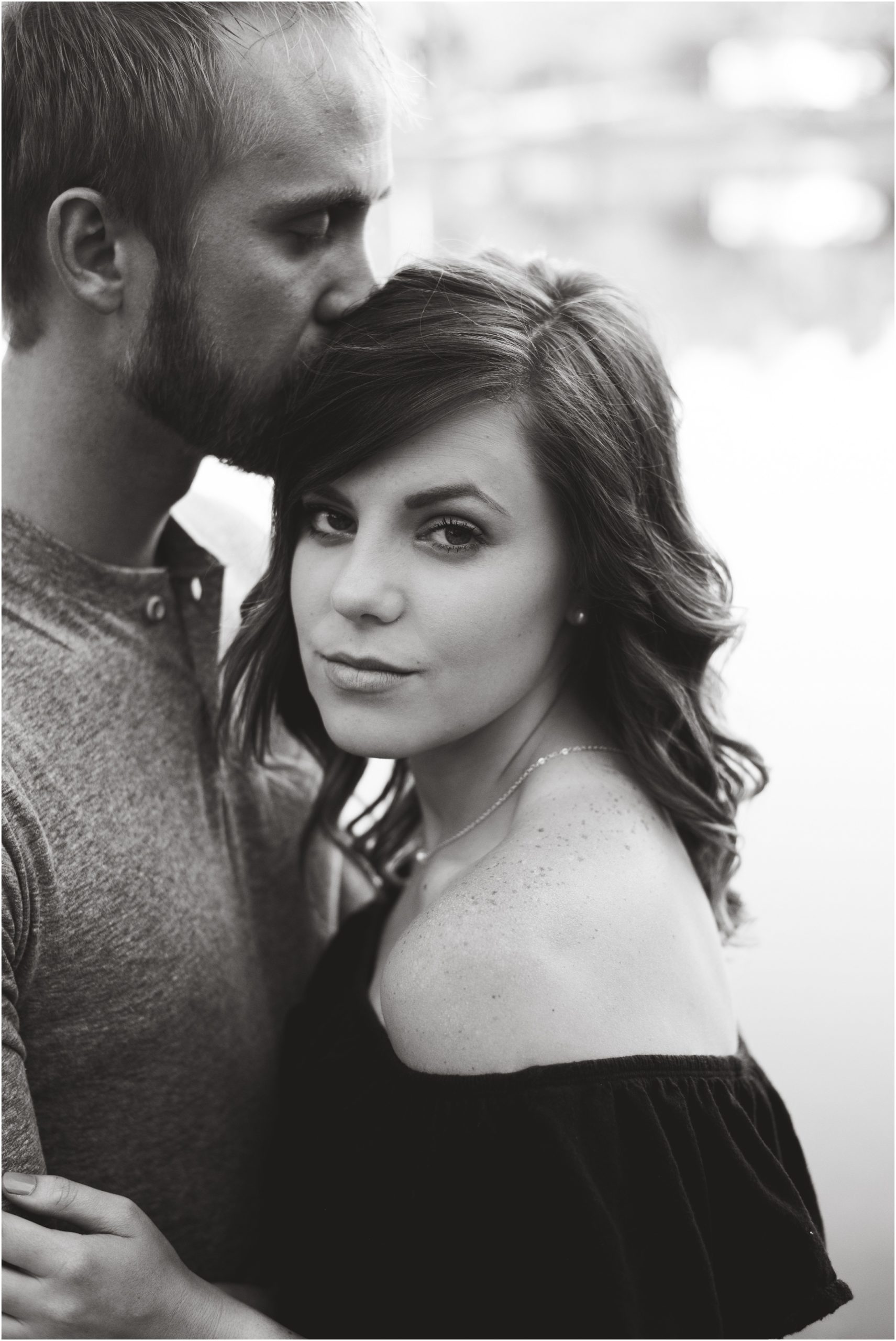 black and white image of girl look at camera while boyfriend kisses head for couples session