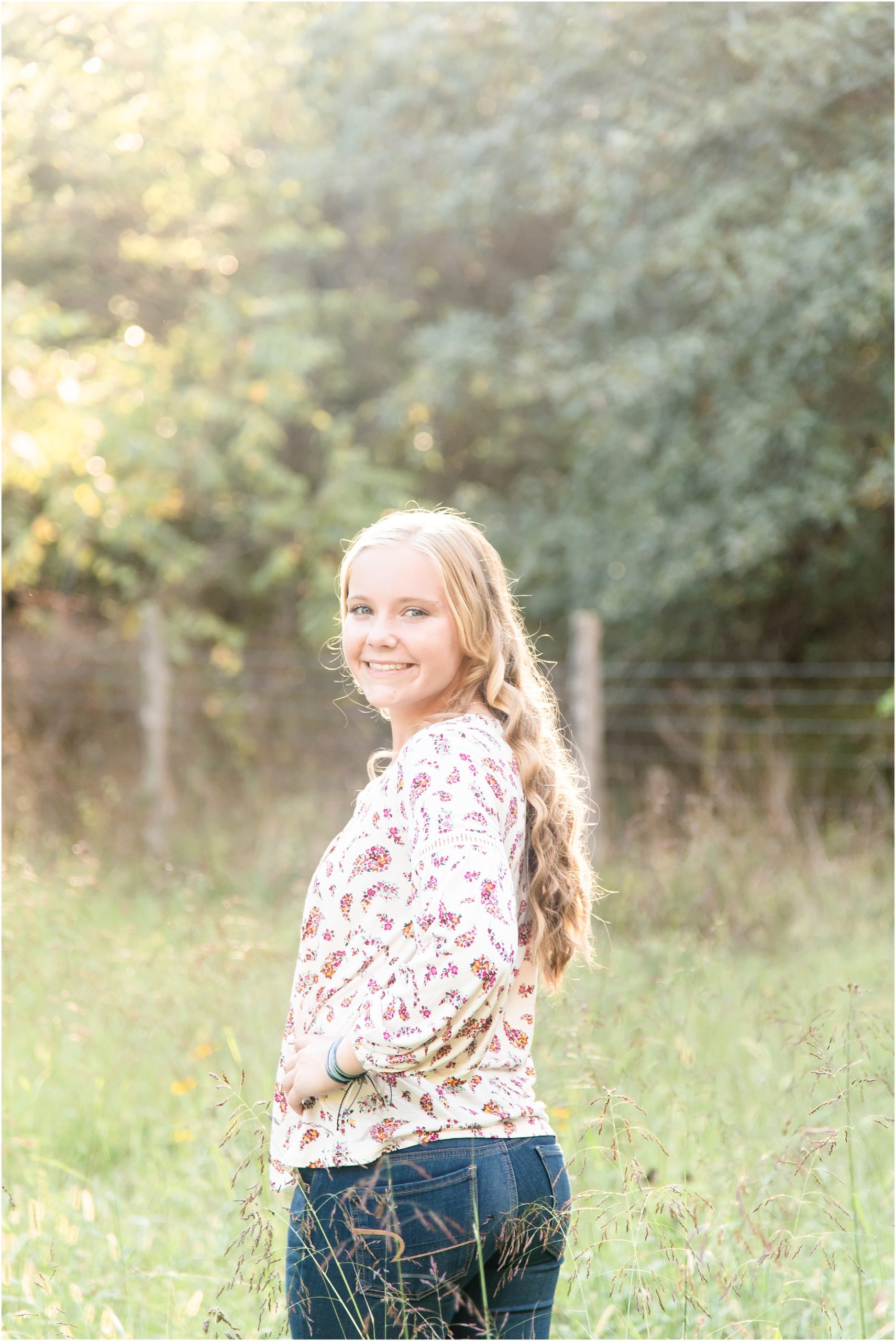 blonde senior girl in floral shirt smiling back at the camera for Russellville high school senior session on family farm in tall grasses and sunlight