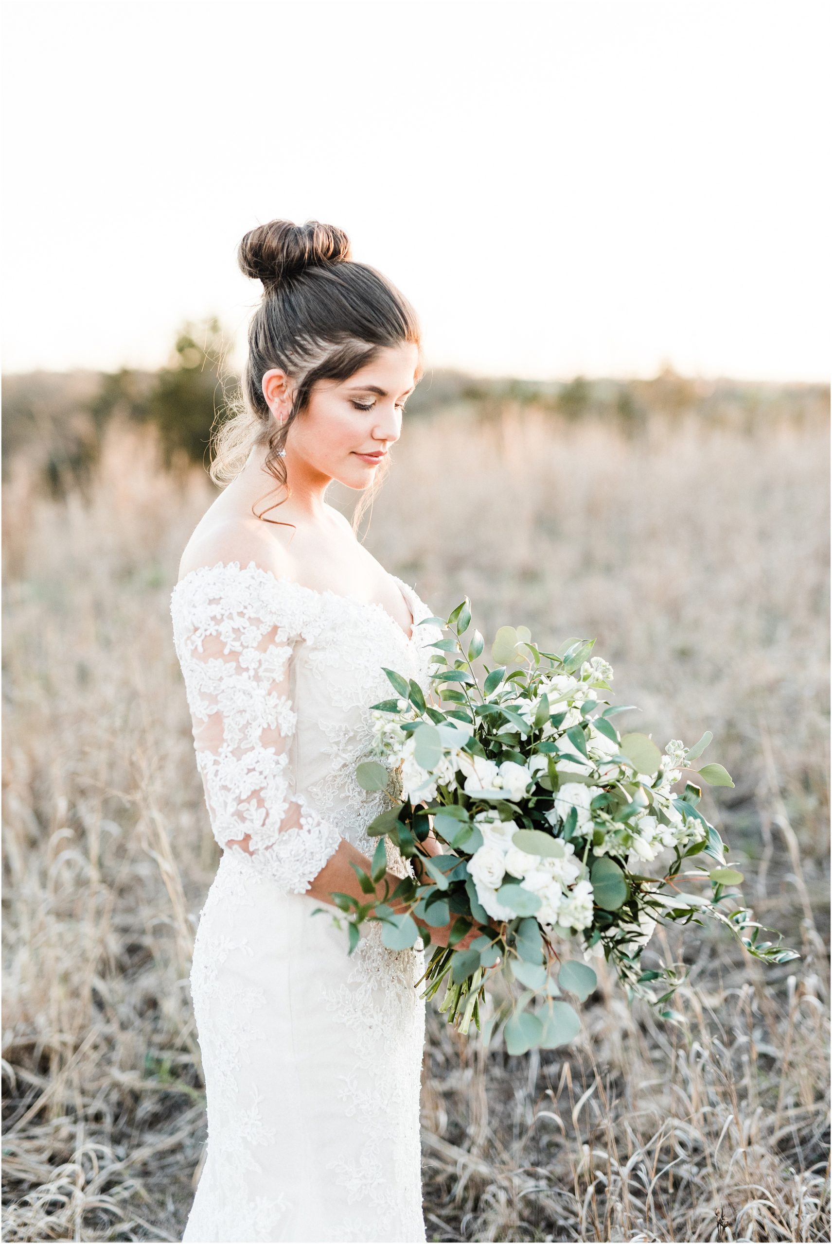 bridal portrait of bride with brown hair up in bun looking down at green and white bouquet during bridal session in field at sunset
