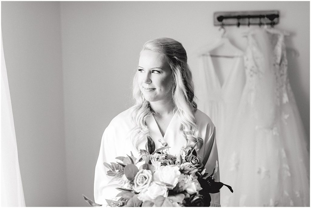 black and white image of bride in robe looking out window with bouquet