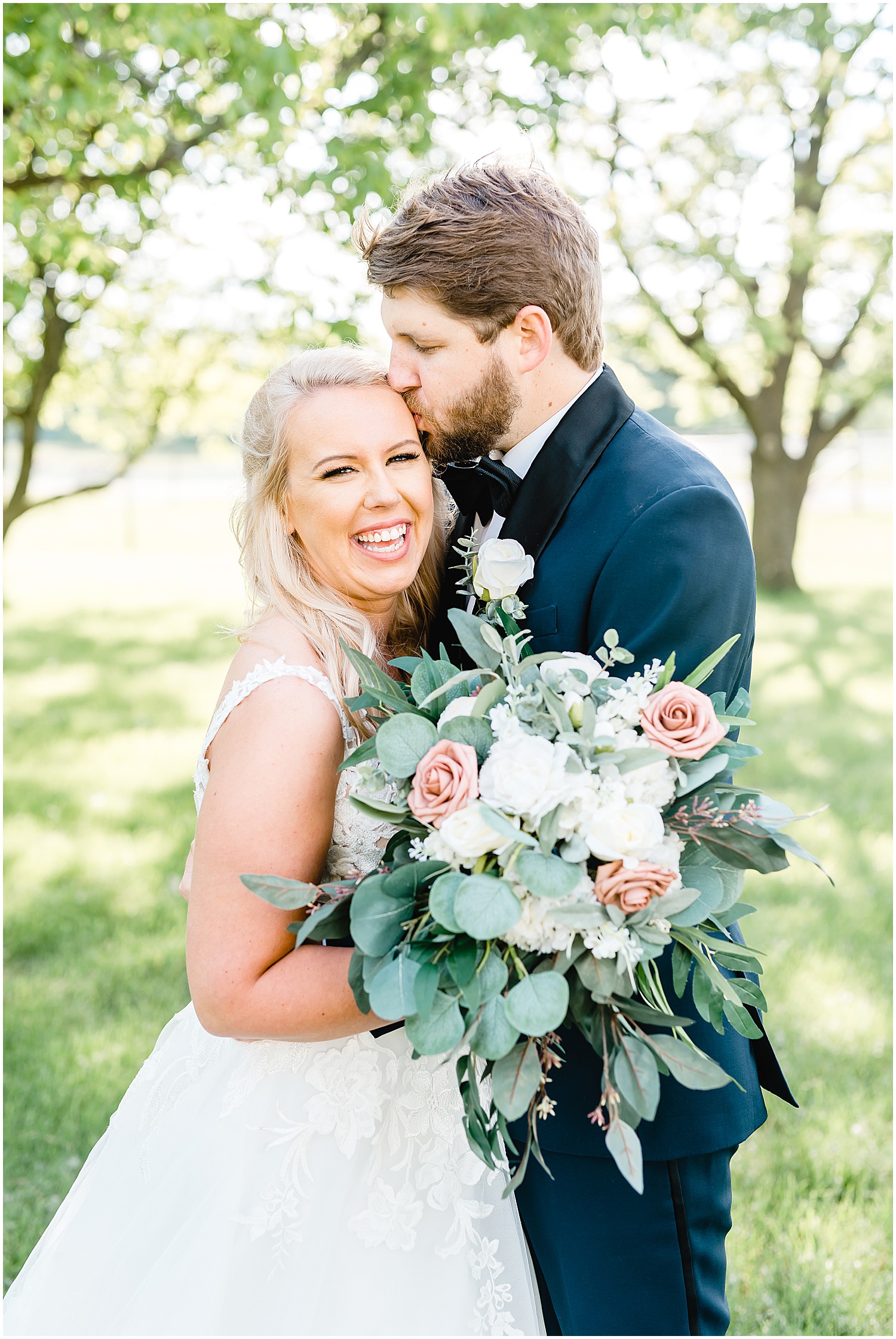 bride smiles at camera with bouquet while groom kisses her forehead