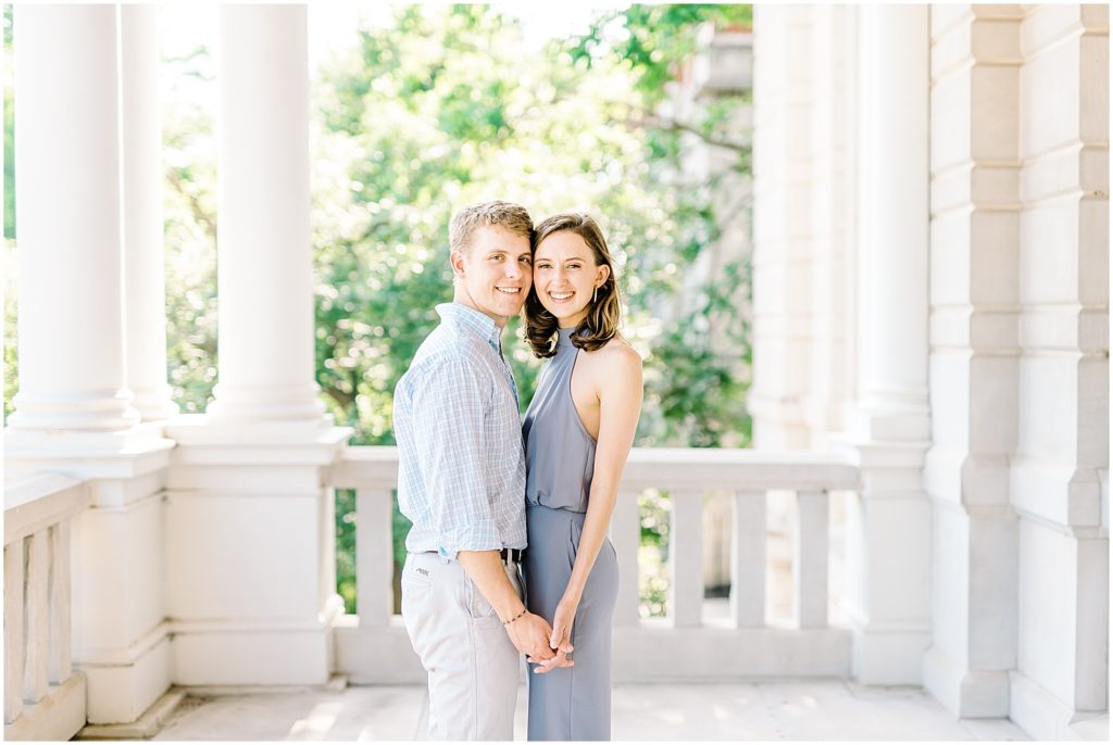 young couple holds hands and smiles while standing on porch on mu campus for engagement session
