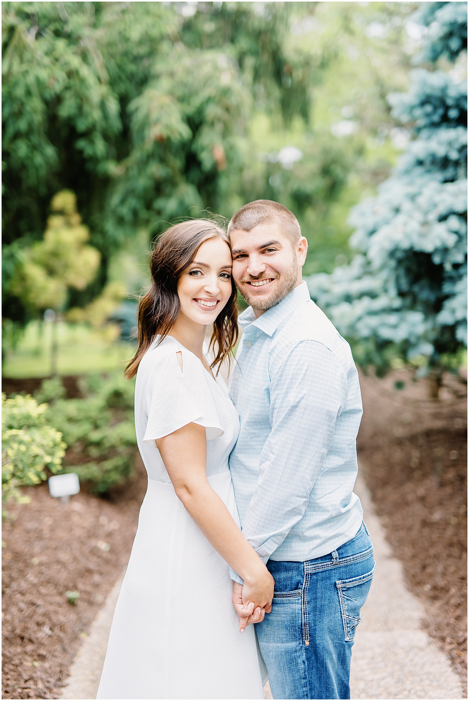couple holding hands and smiling on a sidewalk under trees for engagement session at shelter gardens in columbia, mo