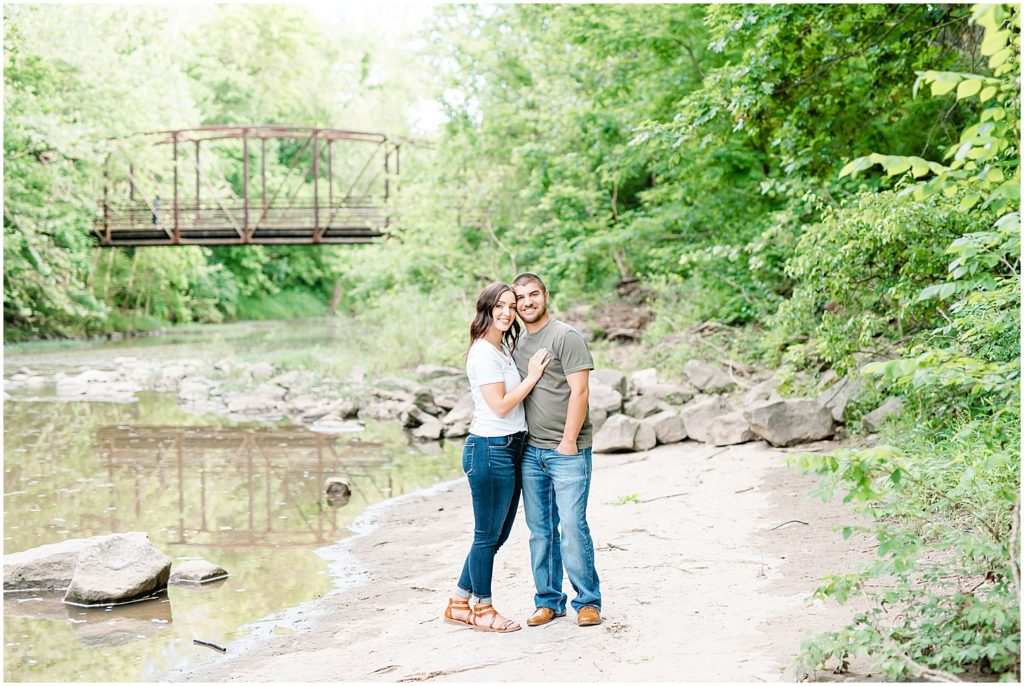 an engaged couple stands together in a creek bed with a bridge in the background at capen park for engagement session
