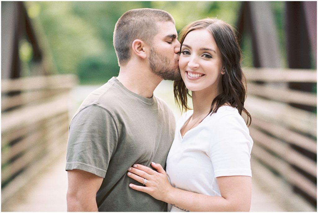 a woman smiles at the camera while her fiance kisses her on the cheek on a bridge at capen park for engagement session