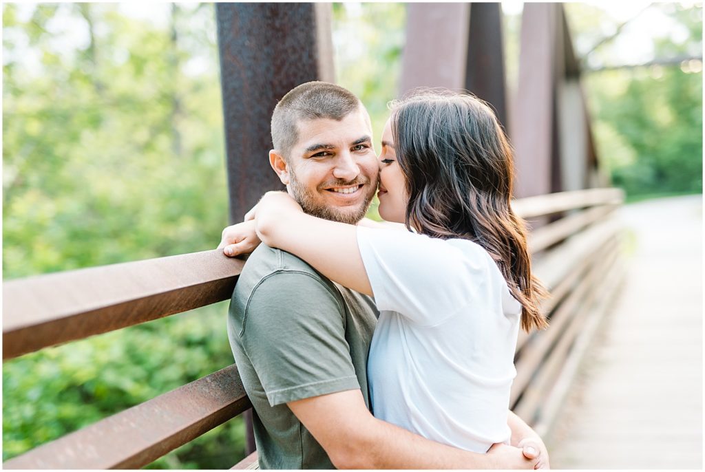a man smiles at the camera as his fiance kisses him on the cheek on a bridge for capen park engagement session