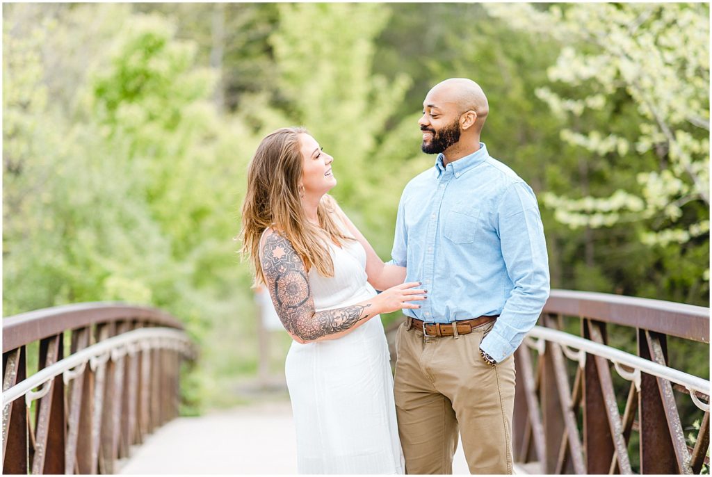 engaged couple laughs on bridge with green trees in background