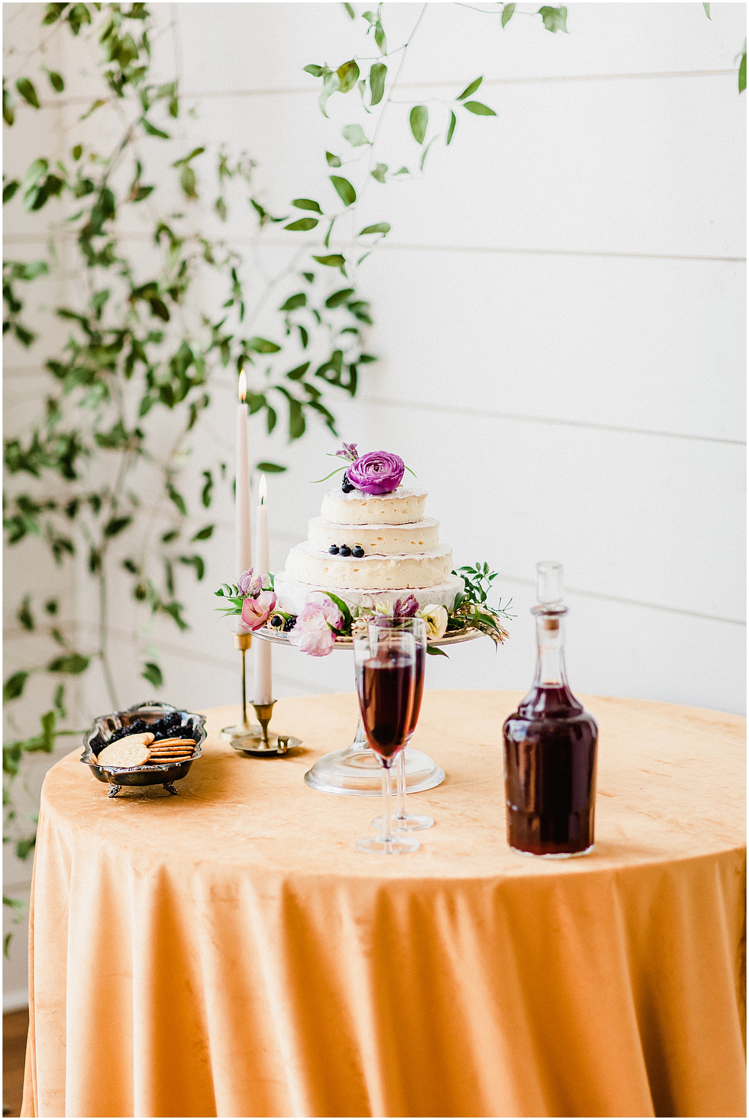 a brie cake featuring blueberries and purple flowers sits on a velvet covered table with wine and crackers
