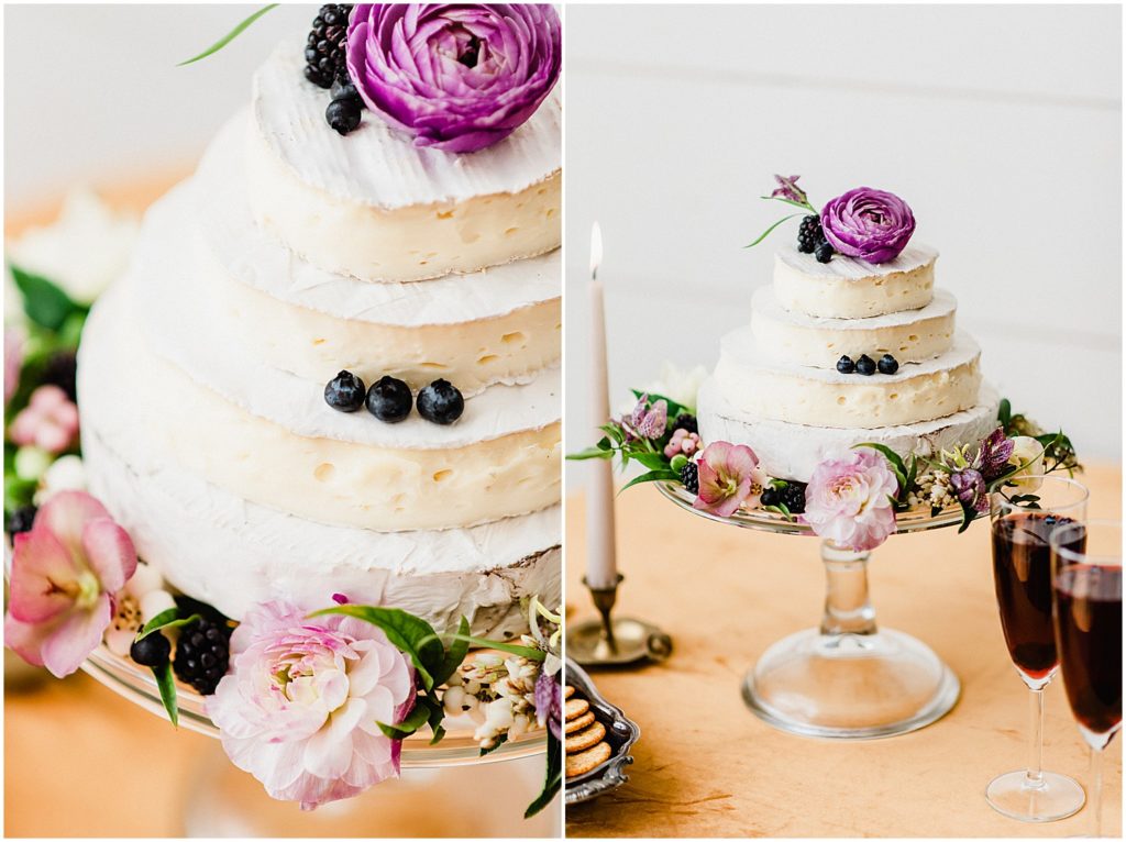 a brie cake featuring blueberries and purple flowers sits upon a linen covered table and glass cake stand