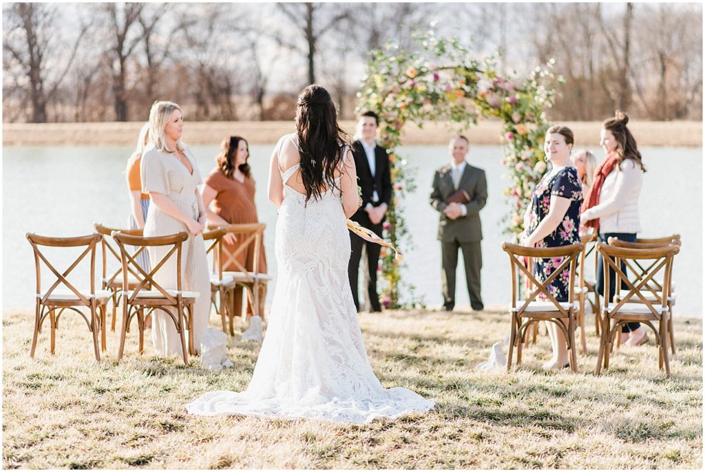 a bride stands at the top of the wedding aisle looking out over a large lake and floral covered arch