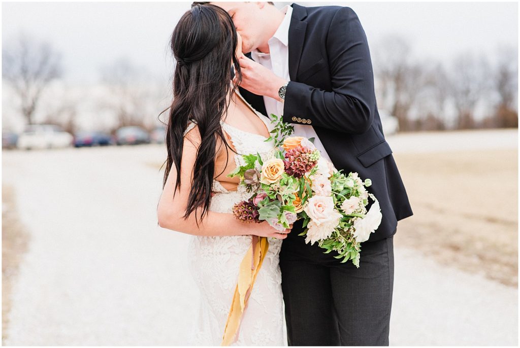 a bride and groom kiss on the driveway outside of Emerson Fields venue holding a large bouquet of flowers