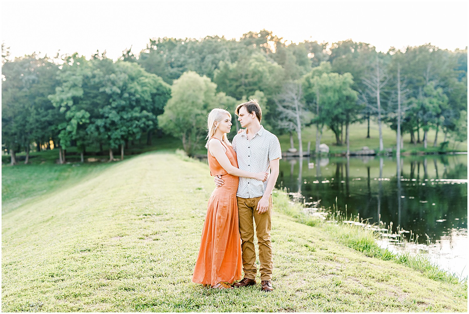 engaged couple stands together on grassy banks of lake wearing fall colors