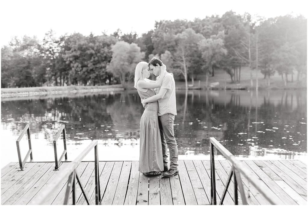 black and white image of young couple holding each other on dock of a lake at kempkers back 40 wedding venue in missouri for engagement session