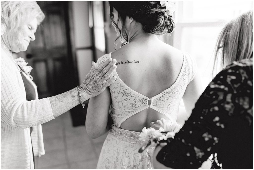 black and white image of grandma patting shoulder of bride getting into dress next to window
