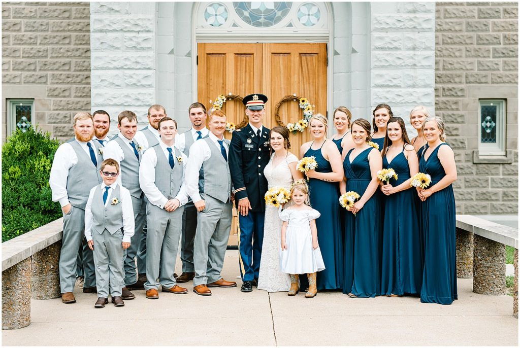 full wedding party in gray suits and navy dresses standing with bride and groom in front of church on wedding day