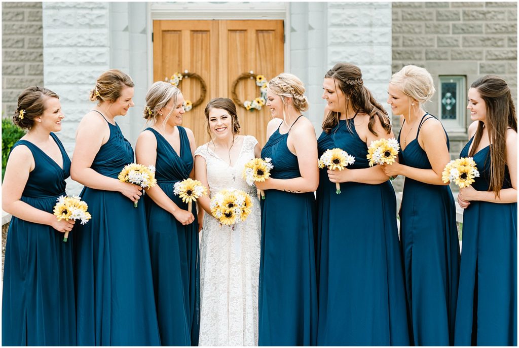 bride and bridesmaids in long navy dresses smile for wedding party pictures outside vienna, mo church