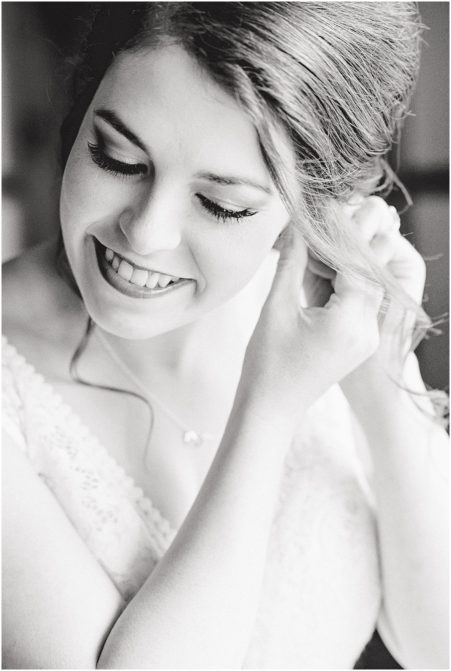 black and white image of bride putting earring on before wedding next to window