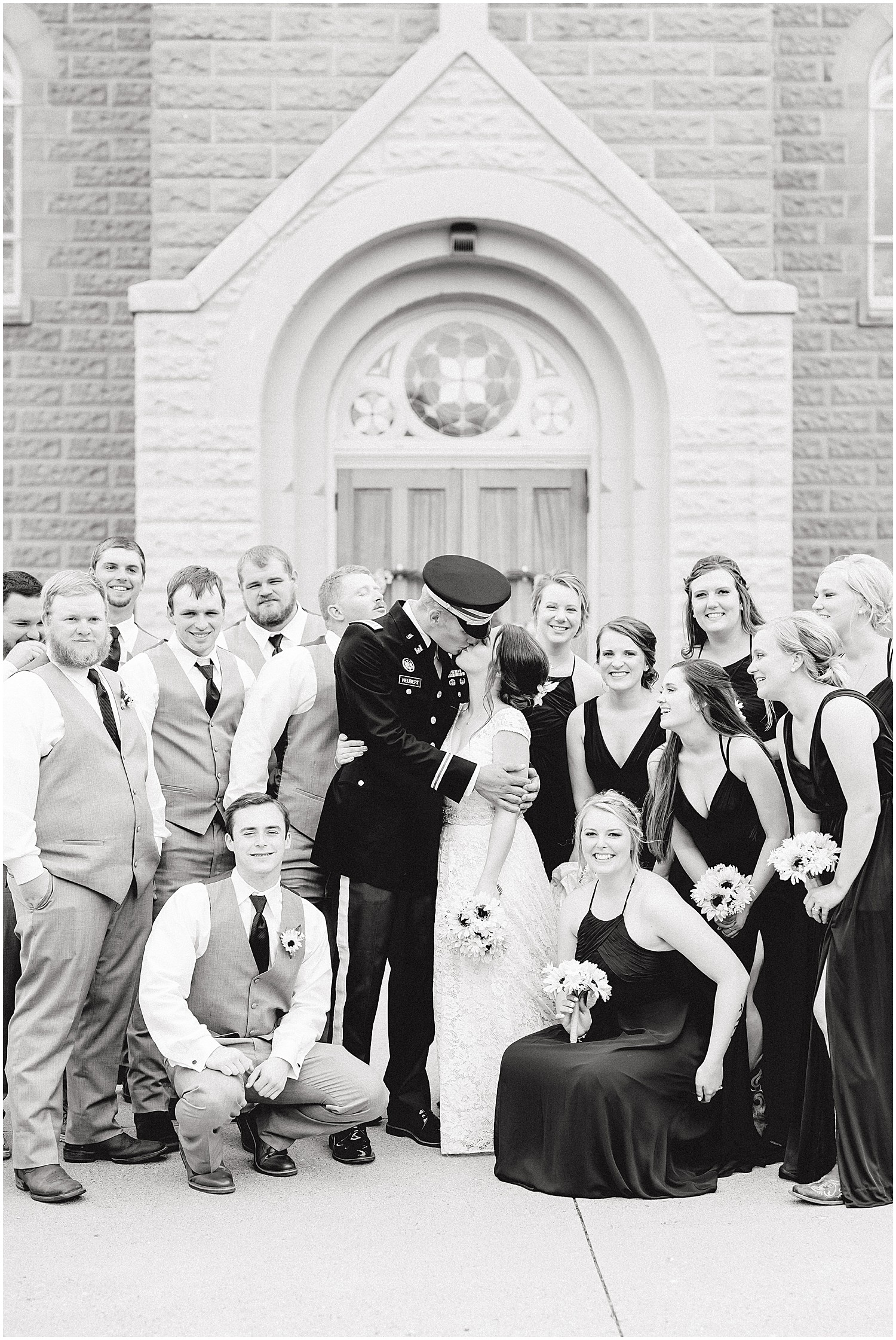 black and white image of bride and groom kissing surrounded by wedding party in front of church