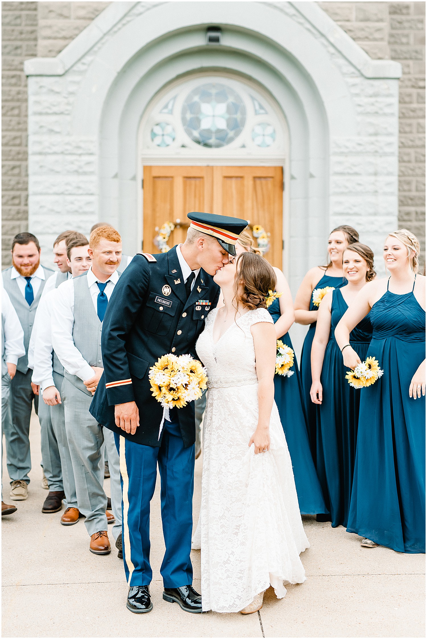 bride and groom kiss outside church with wedding party in the background in gray suits and long navy dresses