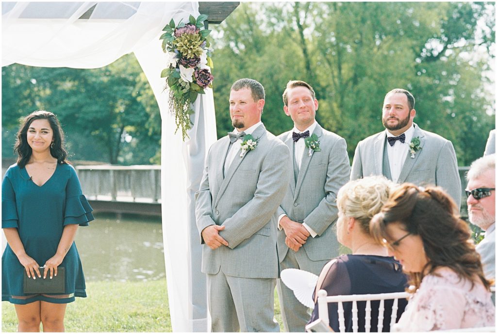 a groom stands at the altar waiting for his bride to walk down the aisle with bridge and lake in the background