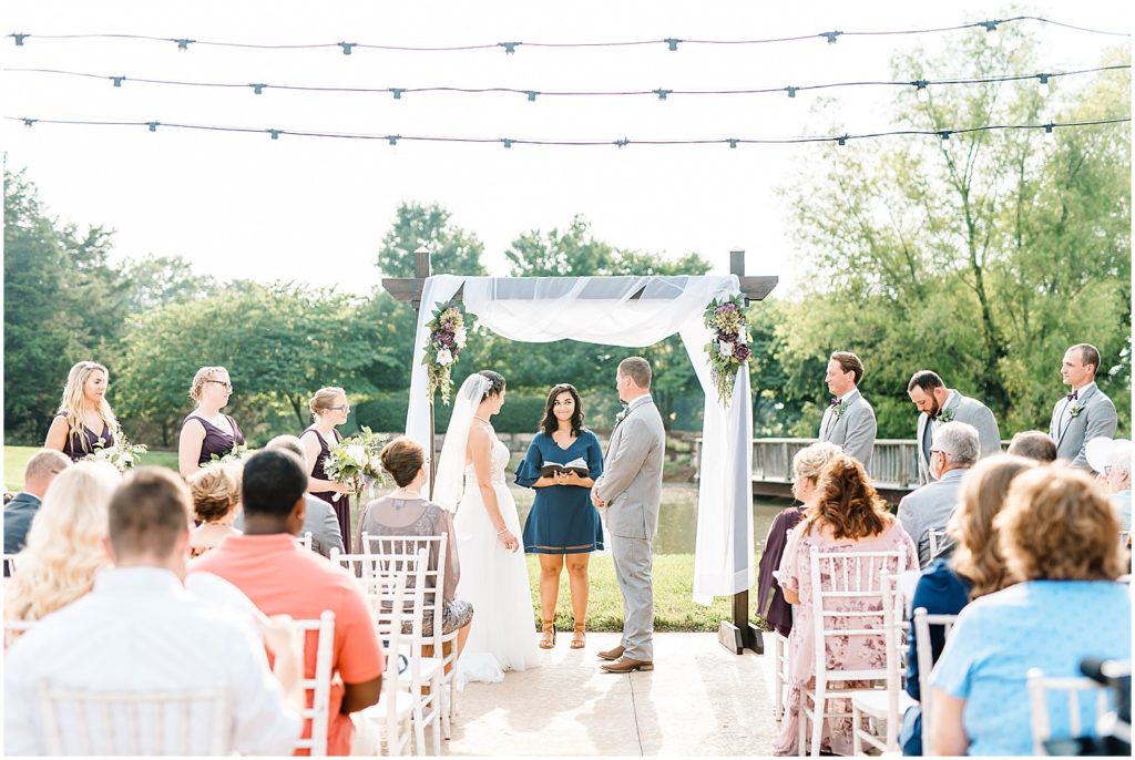 wedding party stands at the front of wedding ceremony with wood arbor draped with white curtain and purple flower arrangements