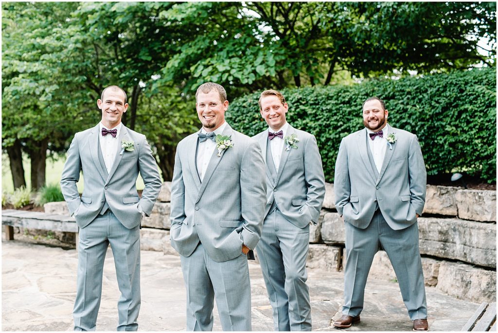 groom and groomsmen in gray suits and purple bow ties smile at the camera