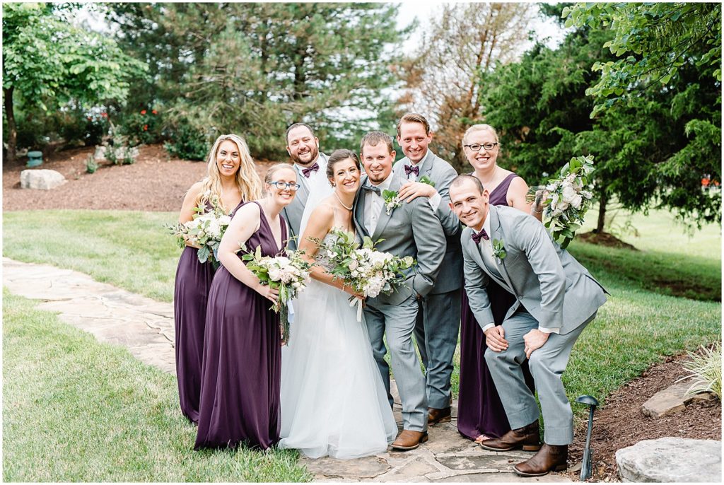wedding party smiles at the camera surrounding bride and groom during cedar lake cellars wedding in wright city, mo