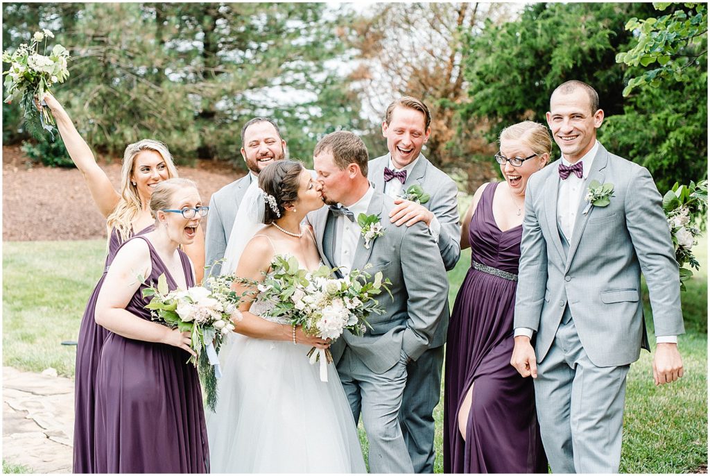 bride and groom kiss as wedding party laughs during portraits at cedar lake cellars wedding