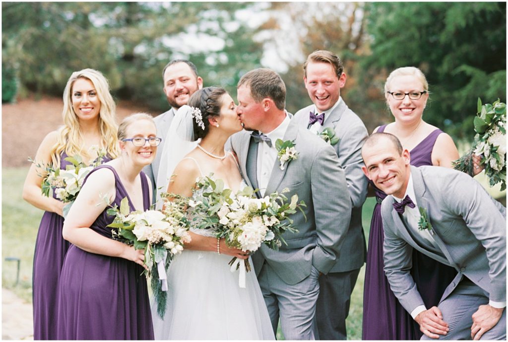wedding party in gray and purple surround bride and groom as they kiss during portraits at cedar lake cellars wedding